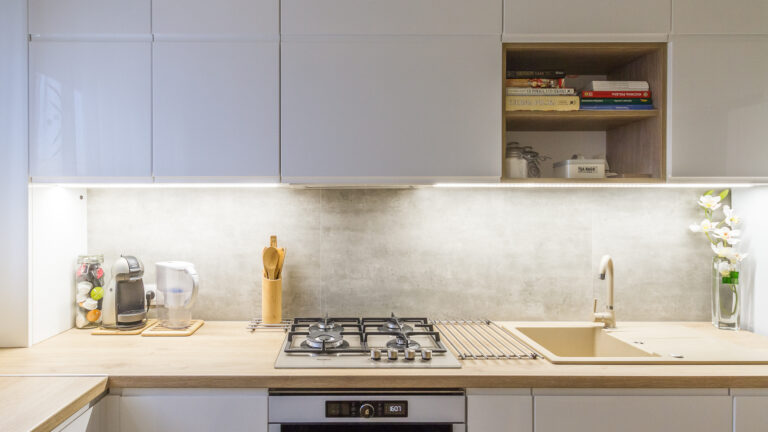 Nowoczesna kuchnia z białymi szafkami, drewnianym blatem, płytą gazową i beżowym zlewozmywakiem. Na blacie ekspres do kawy, dzbanek filtrujący oraz drewniany pojemnik na łyżki, obok wazon z białymi kwiatami.